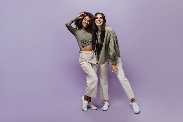 Chicas con estilo positivo con peinado morena en pantalones beige, zapatillas blancas y camisas verde oliva sonriendo y mirando a la cámara