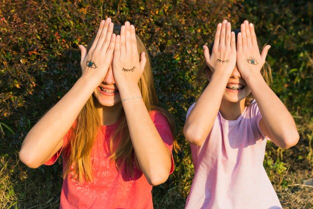 Chicas felices de pie delante de la planta cubriendo sus ojos con tatuaje en la mano