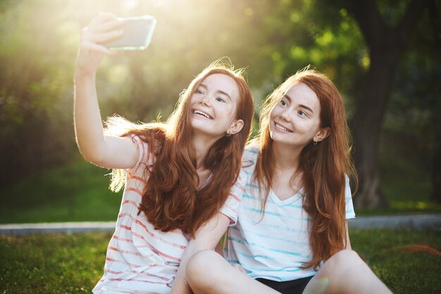 Chicas gemelas de jengibre tomando un selfie en un teléfono inteligente, sonriendo con regocijo. La tecnología moderna conecta a las personas más que nunca. Tener un amigo lejano es muy divertido.