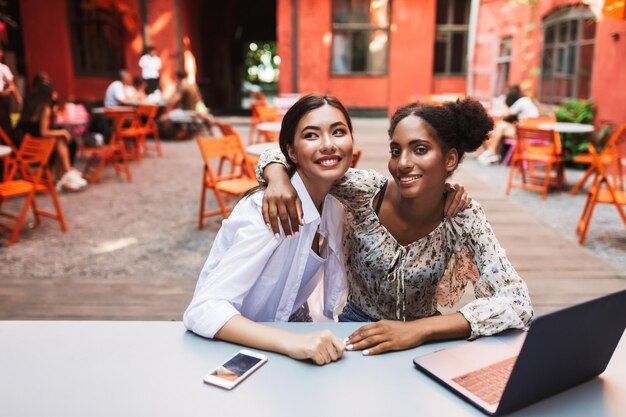 Chicas guapas amistosas abrazándose mientras trabajan felizmente con una laptop en el acogedor patio de la cafetería