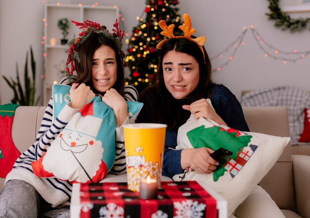 Chicas guapas asustadas con corona de acebo y diadema de renos sostén almohadas y miran televisión sentados en sillones Navidad en casa