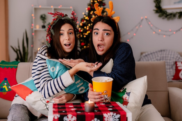 Chicas guapas asustadas con corona de acebo y diadema de renos sostienen palomitas de maíz y miran televisión sentados en sillones Navidad en casa
