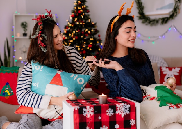 Chicas guapas complacidas con corona de acebo y diadema de renos sostienen adornos de bolas de vidrio y teléfono sentados en sillones y disfrutando de la Navidad en casa