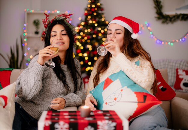 Chicas guapas complacidas con sombrero de santa beben copas de champán sentados en sillones y disfrutando de la Navidad en casa