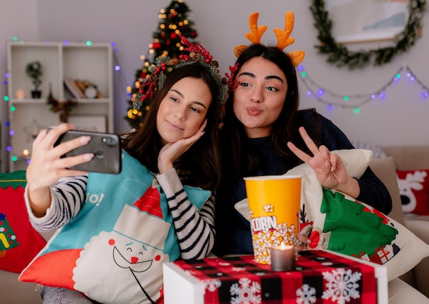 Chicas guapas contentas con corona de acebo y diadema de renos toman selfie mirando el teléfono sentados en sillones y disfrutando de la Navidad en casa