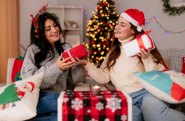 Chicas guapas contentas con gorro de Papá Noel y corona de acebo, sostén y miran cajas de regalo sentadas en sillones y disfrutando de la Navidad en casa