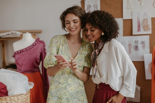 Las chicas guapas miran la pantalla del teléfono Las damas con elegantes trajes de verano sonríen y posan en la acogedora oficina del diseñador de moda