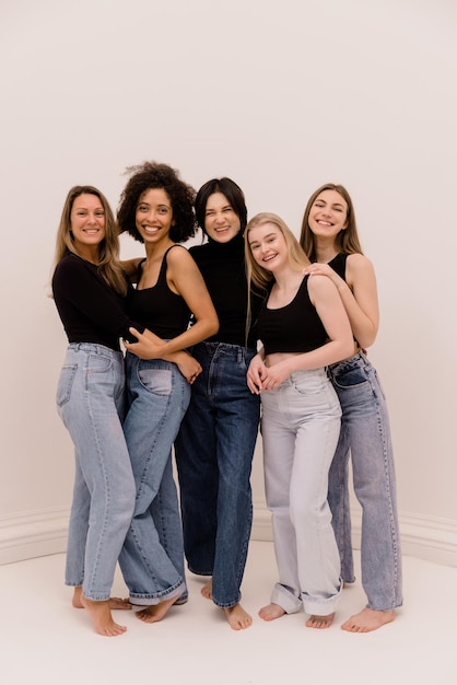 Chicas interraciales jóvenes y adultas felices de longitud completa en sudaderas negras y jeans miran a la cámara sobre fondo blanco