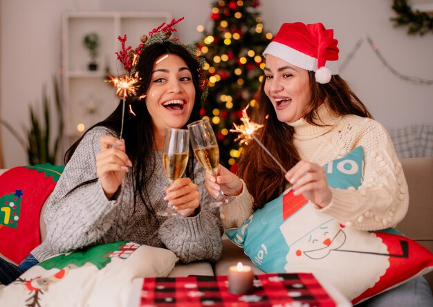 Chicas jóvenes bonitas alegres con gorro de Papá Noel sostienen copas de champán y bengalas sentados en sillones y disfrutando de la Navidad en casa