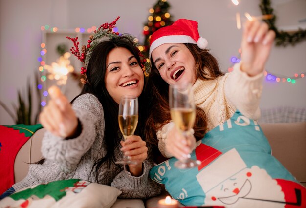 Chicas jóvenes bonitas alegres con gorro de Papá Noel sostienen copas de champán y bengalas sentados en sillones y disfrutando de la Navidad en casa