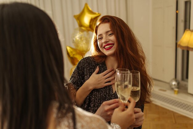 Chicas jóvenes felices en vestidos levantando copas brindando por el cumpleaños de un amigo pelirrojo en la habitación concepto de emociones navideñas