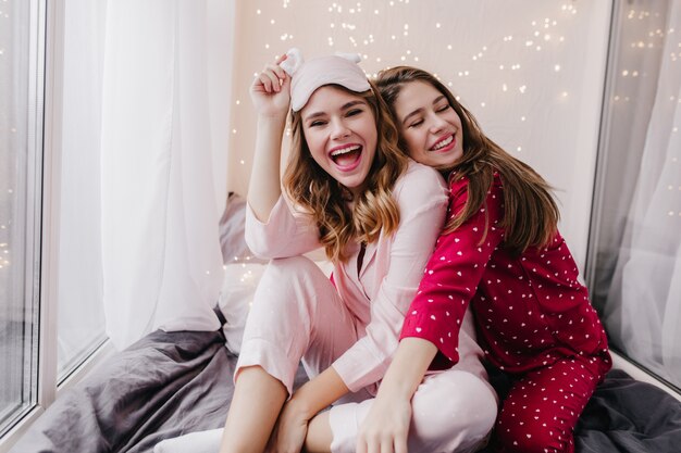 Chicas maravillosas en pijamas lindos sentados en una sábana oscura. Señoras encantadoras caucásicas que se divierten juntos en la mañana.