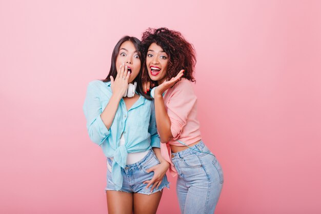 Chicas morenas alegres en atuendos casuales de moda posando con expresión de cara de sorpresa. Foto interior de adorables señoritas con cabello negro de pie en la habitación rosa.