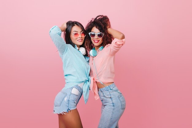 Chicas morenas entusiastas jugando con el pelo mientras posan, descansando juntos. Alegre mujer de pelo oscuro en pantalones cortos de jeans bailando con un amigo africano.