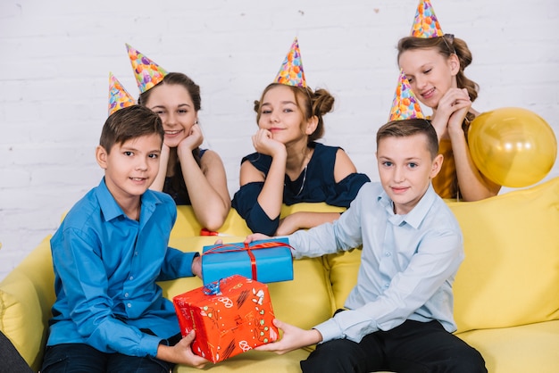 Chicas de pie detrás del sofá mirando a dos adolescentes sosteniendo cajas de regalo