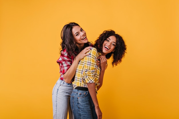 Las chicas positivas se relajan y se divierten en la sesión de fotos en la habitación amarilla. Retrato de risa a chicas bronceadas con pelo rizado.