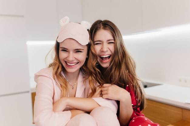 Chicas románticas con cabello brillante riendo mientras están sentadas en la cocina. Maravillosas hermanas caucásicas en pijama posando con placer en la sala de luz.