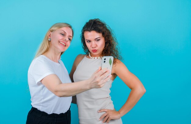 Las chicas rubias y morenas se están tomando selfie con el móvil en el fondo azul