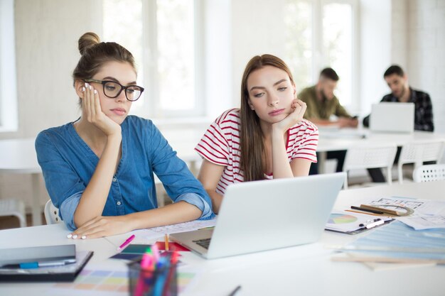 Chicas serias apoyándose en las manos mientras trabajan cuidadosamente con una computadora portátil Las mujeres jóvenes pasan tiempo juntas en la oficina moderna