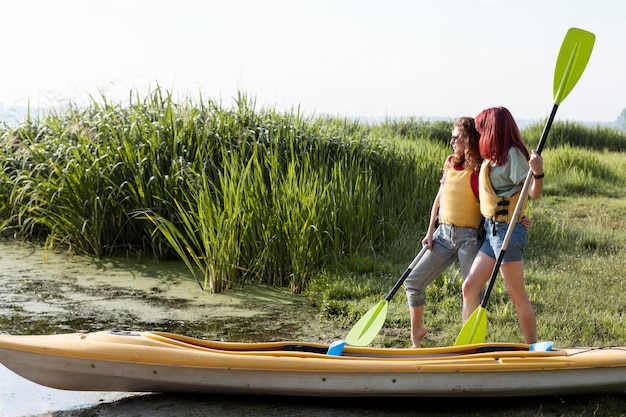 Chicas de tiro completo de pie cerca de kayak sosteniendo remos