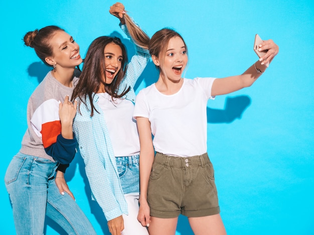 Chicas tomando fotos de autorretrato autofoto en smartphone.Modelos posando cerca de la pared azul en el estudio, hembra mostrando emociones positivas