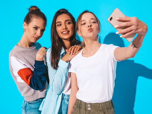 Chicas tomando fotos de autorretrato en el teléfono inteligente. Modelos posando cerca de la pared azul en el estudio. Mujer haciendo cara de pato en la cámara frontal