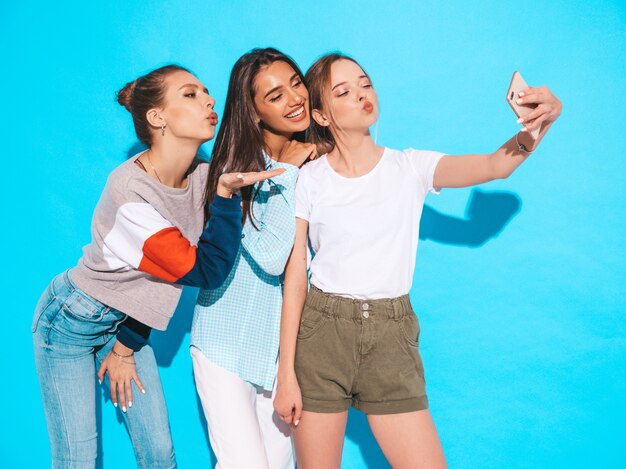 Chicas tomando fotos de autorretrato en el teléfono inteligente. Modelos posando junto a la pared azul en el estudio. Mujeres mostrando emociones positivas. Dan un beso al aire