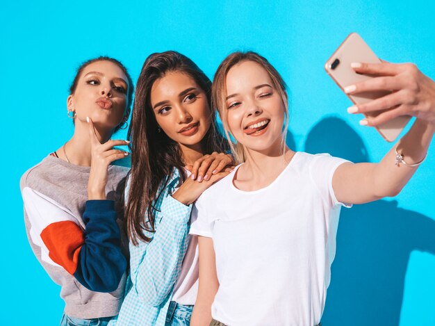 Chicas tomando fotos de autorretrato en el teléfono inteligente. Modelos posando junto a la pared azul en el estudio. Mujeres mostrando emociones positivas