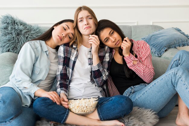 Chicas tristes sentadas en el sofá y comiendo palomitas y haciendo muecas