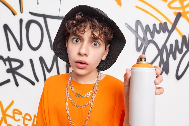 Un chico adolescente sorprendido mira conmocionado a la cámara sostiene una botella de aerosol disfruta creando obras de arte contemporáneo en las paredes de la calle usa ropa de moda lleva el estilo de vida urbano