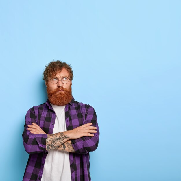 Chico adulto guapo con cabello y barba pelirrojos, cruza los brazos sobre el pecho, tiene una mirada insatisfecha