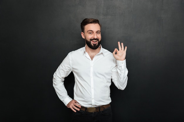 Chico adulto en la oficina posando ante la cámara, sonriendo y gesticulando con un signo OK que expresa que todo está bien sobre gris oscuro