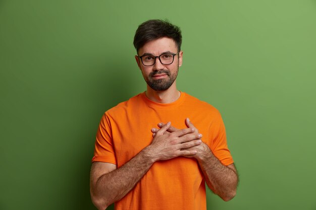 Un chico sin afeitar halagado y complacido presiona las manos al corazón, expresa sentimientos conmovedores y la gratitud aprecia la alabanza, usa gafas transparentes y una camiseta naranja brillante aislada en una pared verde