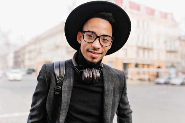 Chico africano encantador con ojos oscuros sonriendo. Retrato al aire libre de joven negro en accesorios casuales posando en la calle.