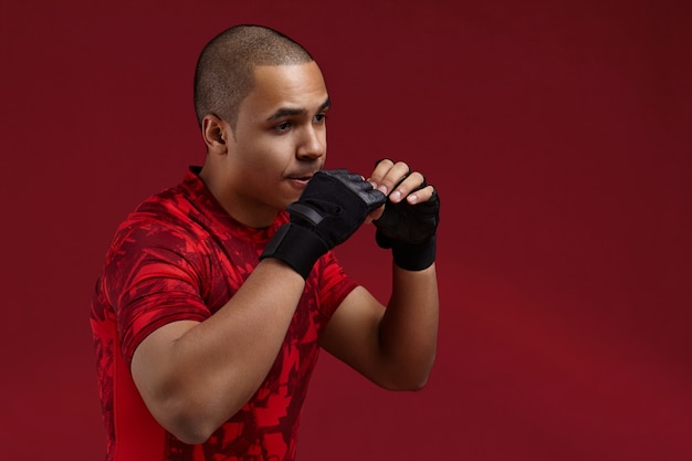 Foto gratuita chico africano guapo con guantes sin dedos de cuero negro entrenando en el gimnasio, trabajando en la técnica de perforación, sintiéndose cansado y agotado. joven luchador de piel oscura con fuertes brazos de boxeo en estudio