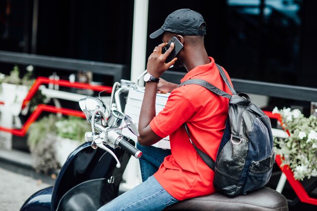 Chico africano joven acepta el pedido por teléfono y escribe en moto sosteniendo cajas con pizza
