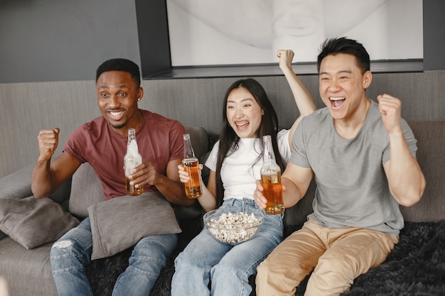 Chico africano y pareja asiática tintinean botellas con una cerveza. Amigos viendo partido de fútbol, comiendo palomitas de maíz. Gente que apoya a un equipo de fútbol.