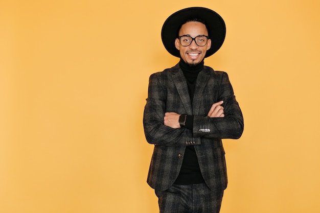 chico africano sonriente con expresión de la cara inspirada. Foto interior del hombre negro complacido con sombrero de pie con los brazos cruzados sobre la pared amarilla.