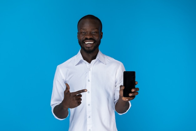 Chico afroamericano barbudo está mostrando celular en camisa blanca