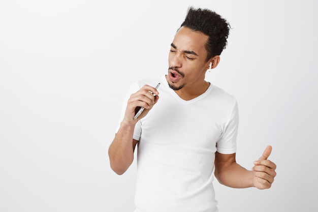 Chico afroamericano despreocupado jugando karaoke, cantando en el micrófono del teléfono inteligente, usando auriculares inalámbricos