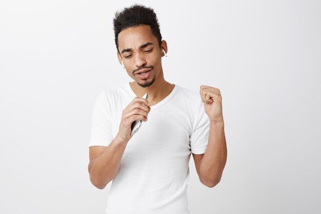 Chico afroamericano despreocupado jugando karaoke, cantando en el micrófono del teléfono inteligente, usando auriculares inalámbricos