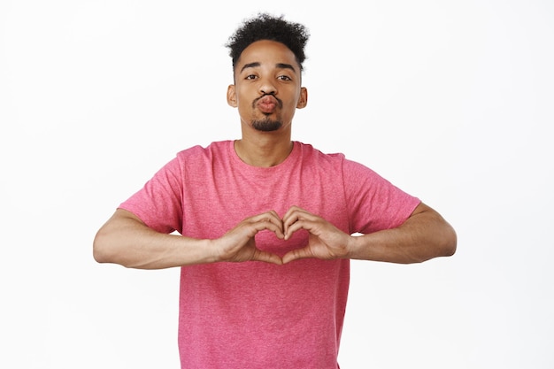 Chico afroamericano encantador y tonto, novio besándose y mostrando un gesto de amor al corazón, te amo cara, de pie contra un fondo blanco con una camiseta rosa.