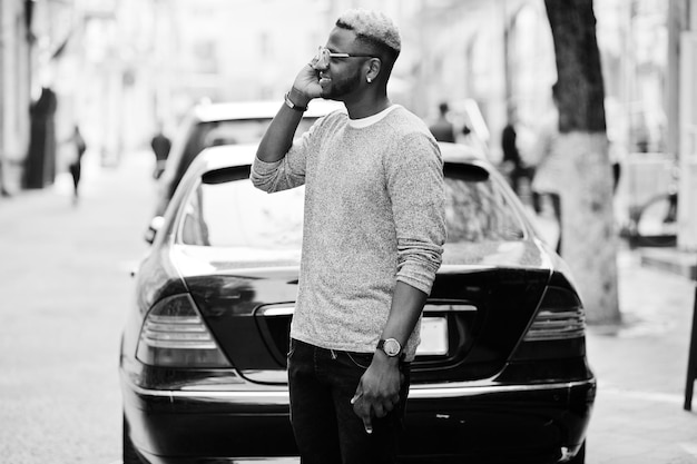 Chico afroamericano con estilo en suéter gris y gafas posó en la calle contra un coche de negocios negro y hablando por teléfono