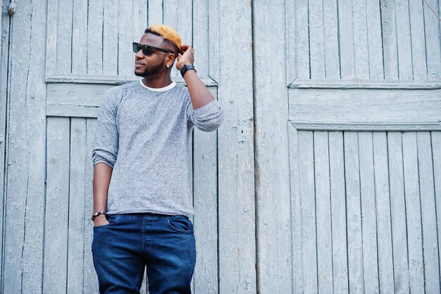 Chico afroamericano con estilo en suéter gris y gafas de sol negras posadas en la calle