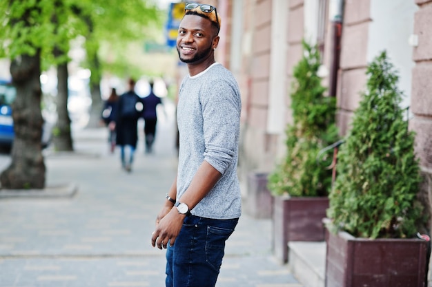 Chico afroamericano con estilo en suéter gris y gafas de sol negras posadas en la calle