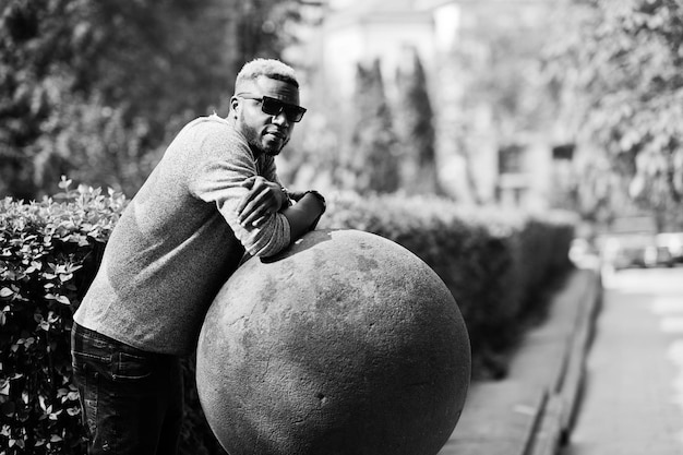 Chico afroamericano con estilo en suéter gris y gafas de sol negras posadas en la calle