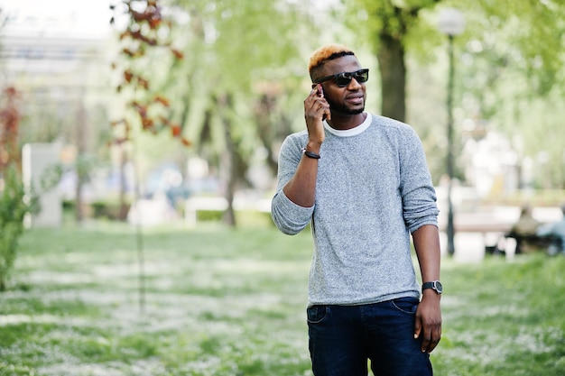 Chico afroamericano con estilo en suéter gris y gafas de sol negras posando en el parque y hablando por teléfono