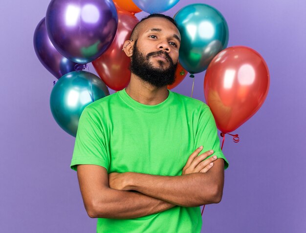 Chico afroamericano joven confiado con camiseta verde de pie delante de globos cruzando las manos aisladas en la pared azul