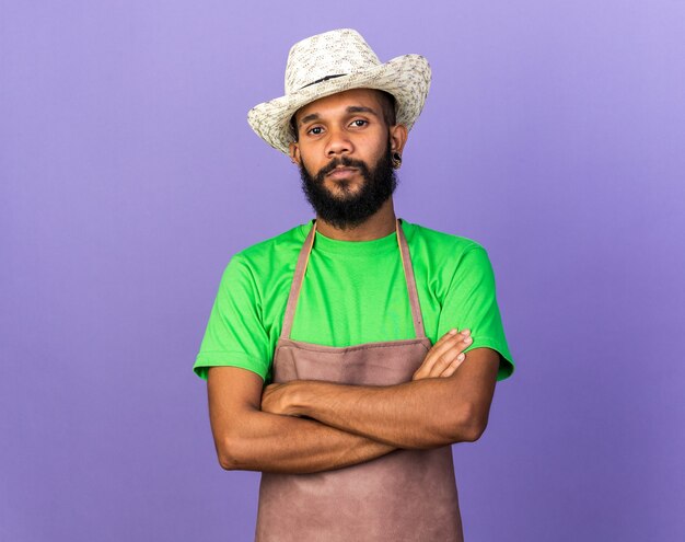 Chico afroamericano joven jardinero confiado con sombrero de jardinería cruzando las manos aisladas en la pared azul