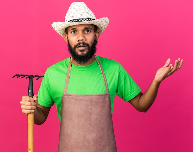 Chico afroamericano joven jardinero confundido con sombrero de jardinería sosteniendo el rastrillo extendiendo la mano